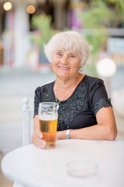 Yazlık bir kafede masada oturan ve uzun bir bardaktan bira içen kıdemli bir kadın.
