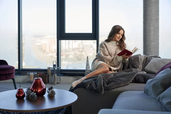 Mulher bonita sentada no sofá e lendo livro interessante — Fotografia de Stock