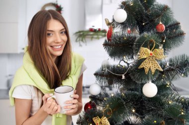 Genç mutlu kadın evde Noel ağacının yanında kahve içiyor..