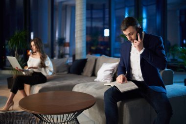 Kadın dizüstü bilgisayar kullanırken, erkek evde telefonla konuşuyor.