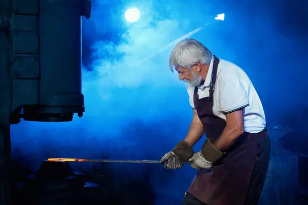 Šedovlasý kovář pracující s roztaveným kovem v kovárně — Stock fotografie