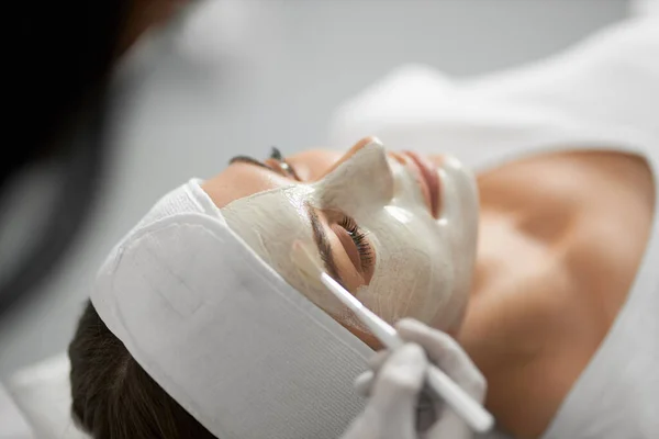 Kosmetička aplikující speciální bílou masku na obličej pro ženu. — Stock fotografie