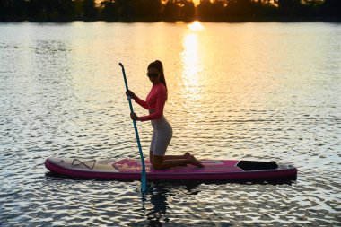 Gün batımında sörf tahtasında sörf yapan zinde genç bir kadın.