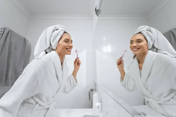 Pretty woman in white robe with towel on head brush teeth in bathroom. — Stock Photo, Image