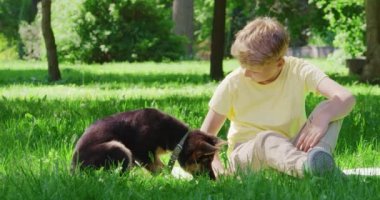 Gülümseyen çocuk çimlerin üzerinde oturuyor ve sevimli köpek yavrusuyla oynuyor.