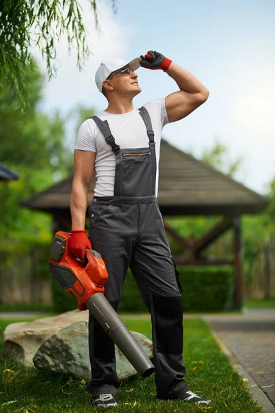 Uomo in tuta da lavoro in piedi in giardino con soffiatore di foglie in mano — Foto Stock