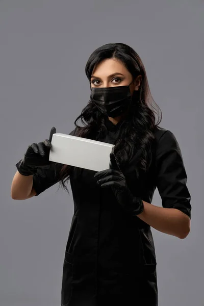 Siyah maskeli ve eldivenli bir kadın kozmetik kutusu tutuyor. — Stok fotoğraf