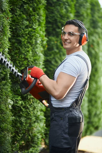Gardener in uniform cutting hedge on back yard — стокове фото