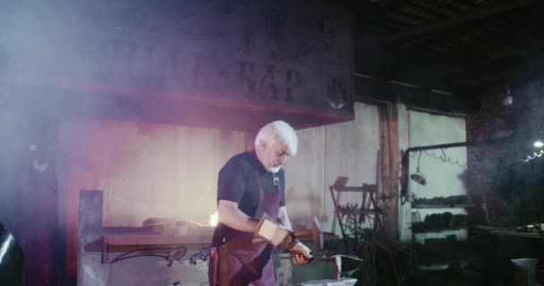 Old blacksmith with grey hair and beard beating with hammer. — Stock video