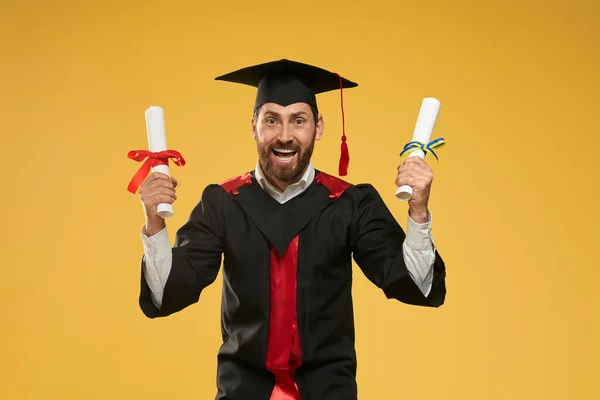 Muž v minometu a maturitní róbě, stojící se dvěma diplomy. — Stock fotografie