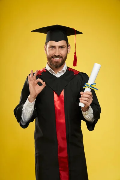 Student wearing graduate gown, mortarboard holding diploma, showing all right. — Fotografia de Stock