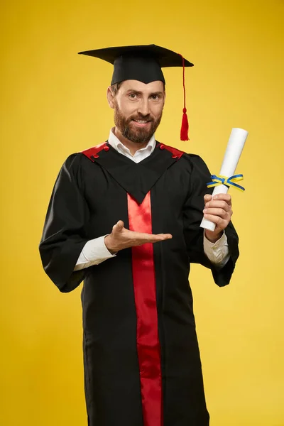 Man wearing graduate gown and mortarboard, showing diploma. — Photo