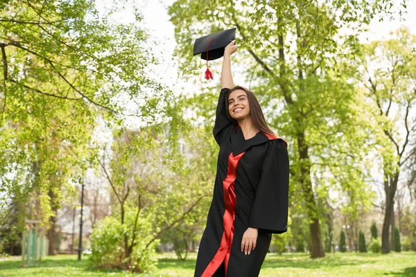 Pretty girl wearing graduate gown raising mortrboard. — Foto Stock