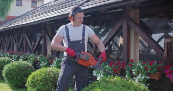 Strong man in earmuffs cutting green bushes with electric machine outdoors. — Vídeo de Stock