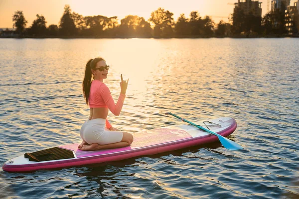 Positive woman sitting on sup board and showing horn gesture — Stockfoto