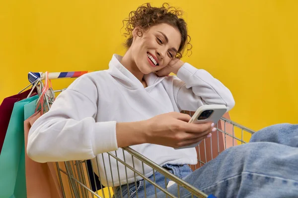 Kaunis nuori nainen istuu ostoskorissa älypuhelimen kanssa. — kuvapankkivalokuva