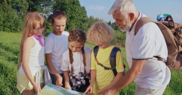 山の中で一緒に地図を保持旅行者のフロントビュー。4人の子供が立って、見て、示す、指して、老人は笑っている。観光と自然発見の概念 — ストック動画