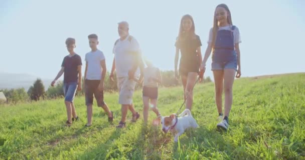 Gri saçlı, Jack Russel Terrier 'ı yöneten ve gülümseyen yaşlı bir adamla ilerleyen bir grup çocuğun ön görüntüsü. Dağlarda birlikte seyahat eden turistler. Takım ruhu kavramı — Stok video