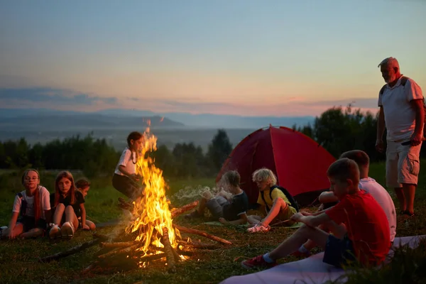 Glückliche Kinder mit erwachsenem Lehrer sitzen abends am Lagerfeuer im Freien. — Stockfoto