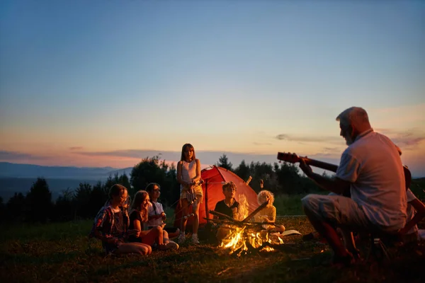 Fröhliche Kinder sitzen am Lagerfeuer und singen in der Abenddämmerung Lieder auf dem Zeltplatz. — Stockfoto