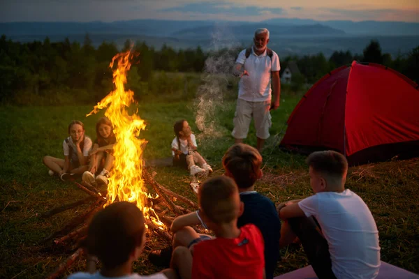 Gruppe von Kindern lauscht Instruktor, während sie in der Dämmerung am Lagerfeuer sitzen. — Stockfoto