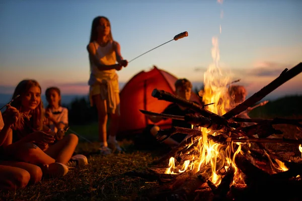 Fröhliche Teenie-Mädchen braten Wurst über dem Feuer, haben Spaß mit Freunden auf dem Campingplatz. — Stockfoto