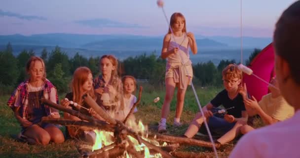 Adolescentes alegres sentados ao redor da fogueira, comendo no acampamento. — Vídeo de Stock