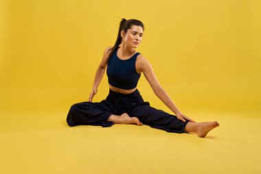 Yoganın bir bacağını öne doğru kıvırıp yere uzanmaya hazırlanan ince bir kız. Esmer kadının Asana 'da yoga yaparken görüntüsü turuncu stüdyo arka planında izole edilmiş. Yoga kavramı.