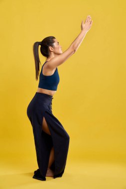 Sıska pantolonlu, sandalyede yoga yapan, geniş bir stüdyoda poz veren zayıf bir kadın. Utkatasana 'da sarı stüdyo arka planında izole edilmiş at kuyruklu Yogi kızı. Yoga asanası kavramı.