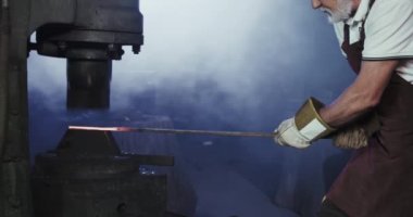 Demircinin zor el işi yaparken yan görüntüsü. Gri saçlı, sakallı, önlük ve eldiven giyen, uzun demir çubuğu olan yaşlı bir adam. Kurnazlık kavramı..