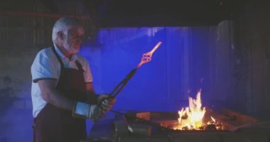 Güçlü demircinin ocakta metal işçiliğini ateşe verişi yan görünüm. Gri saçlı ve sakallı yaşlı adam demircilik, kalpazanlık ve alevler içinde çalışıyor. Demirci sanatı kavramı.