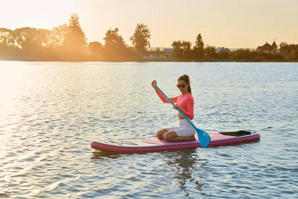 戴着太阳镜 身穿五颜六色运动服的白人女子坐在船舱里 划着长桨划船 水活动和户外运动的概念 — 图库照片