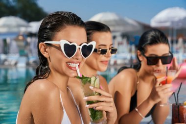 Açık havuzda rahatlayan üç baştan çıkarıcı kızın ön görüntüsü. Sıska, güzel kadınlar suda durup gülümsüyor, konuşuyor, kokteyl içiyor. Dinlenme ve yaz kavramı.