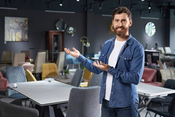 Front view of handsome boy standing, representing furniture in store. Young male with beard looking at camera, smiling, showing furniture, buying, selling. Concept of shopping.