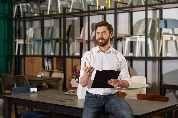 Front view of brunette male with beard sitting on table in furniture store. Handsome designer, architect, seller holding folder, writing, looking aside. Concept of designing.