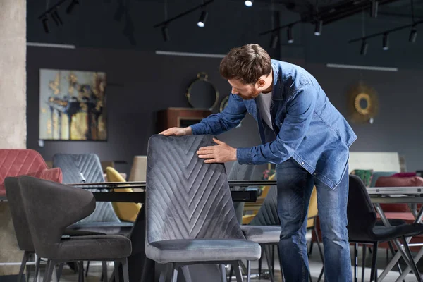 Front view of customer choosing chair in furniture store. Handsome brunette male with beard standing, touching, looking down, buying. Concept of shopping and modern lifestyle.