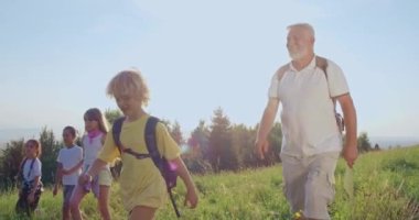 Tepelerde yürüyüş yapan, ileri yürüyen, gülümseyen ve şakalaşan bir grup gezginin ön görüntüsü. Gri saçlı yaşlı adam harita taşıyor, konuşuyor. Seyahat ve doğa keşfi kavramı.