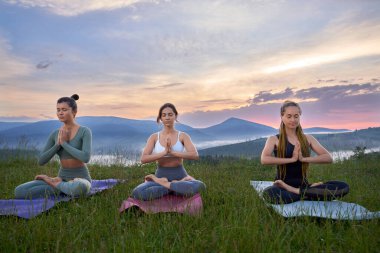 Spor kıyafetli üç genç kadın nilüfer çiçekli yoga minderinde oturup gözleri kapalı meditasyon yapıyorlar. Arka planda dağların ardında şaşırtıcı yaz günbatımı.