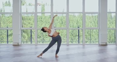 Beyaz spor sutyen ve gri taytlı beyaz kadın geniş eğitim salonunda yoga egzersizleri yapıyor. Arka planda panoramik pencereler olmasına rağmen dağlarda şaşırtıcı manzara.
