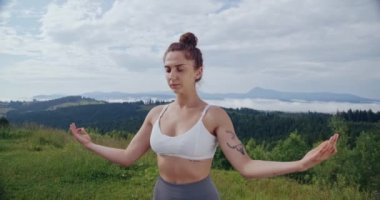 Güzel dağların yaz doğası arasında meditasyon yapan genç bir kadın. Vücudunda dövmesi olan ve beyaz spor sutyen ve gri taytlı bir bayan sporcu..