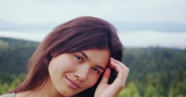 Koyu renk saçlı kadın, dağların arasında doğada dikilirken kameraya bakıyor. Spor elbiseli genç bir kadın dışarıda antrenman yapmak için boş zaman harcıyor..