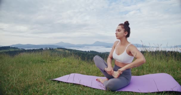 Zdravá fit žena ve sportovní podprsenka a legging sedí na podložce jógy a protahuje nohy. Aktivní žena cvičící jógu pózuje na čerstvém vzduchu.
