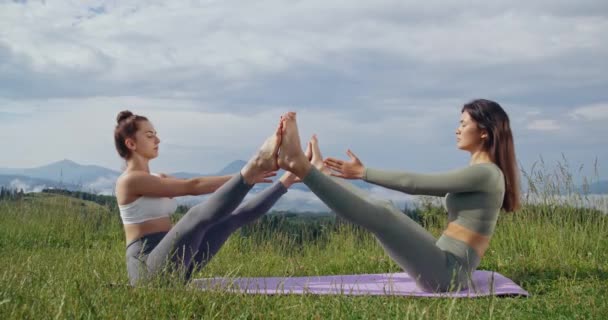 スポーツウェアの若い女性選手は屋外でフィットネス運動をストレッチします 背景に美しい緑の山々 瞑想とバランスの概念 — ストック動画