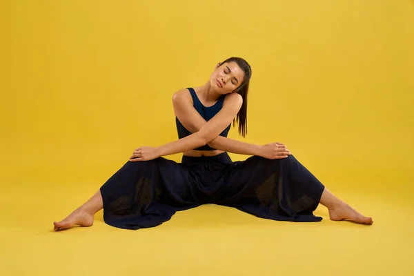 Young woman doing stretching exercise, sitting with legs wide apart indoor. Front view of relaxed girl in black, with crossed arms on knees, isolated on orange background. Concept of meditation.