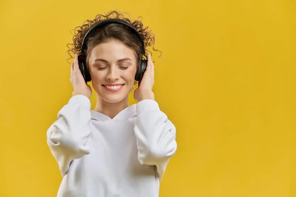 Joyful woman with modern earphones listening music, having fun indoor. Portrait view of female music lover enjoying sound, with closed eyes, isolated on orange studio background. Concept of enjoyment.