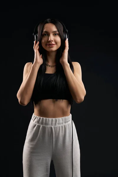 Happy girl in crop top, looking at camera, while listening to music inside. Portrait view of slim female holding headphones, enjoying sound, isolated on black studio background. Concept of music fan.