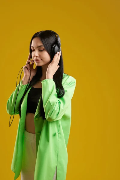 Relaxed woman with closed eyes standing and listening to music indoor. Side view of focused pretty girl wearing headphones, enjoying music, isolated on orange studio background. Concept of music fan.