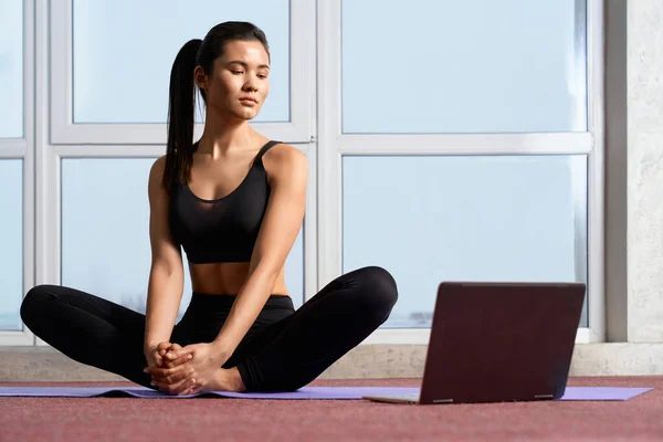 Front view of beautiful yoga instructor streaming online yoga class, using a laptop. Flexible female yogi meditating in butterfly position, while teaching yoga interactive at home. Concept of yoga.