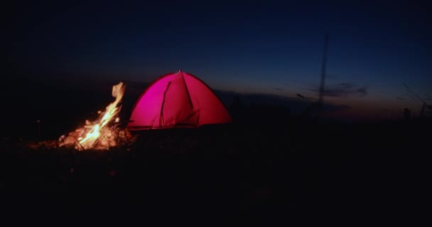 在暮色的群山中 红色帐篷和黑暗天空下篝火的前景 篝火熊熊燃烧的营地 在黄昏时发出火花 并在背景上搭上旅游帐篷 旅行的概念 — 图库视频影像