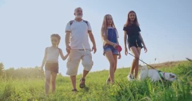 Yazın yeşil tepe boyunca yürüyen teriyerli erkek ve kadın çocukların ön görüntüsü. Üç kızı ve köpeği kırsalda birlikte vakit geçiren mutlu orta yaşlı bir baba. Aile gezisi kavramı.